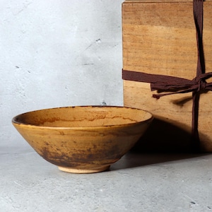 May include: A handmade ceramic bowl with a wide, shallow design. The bowl has a warm, earthy colour palette with brown and tan tones. A wooden box tied with a brown ribbon is in the background.