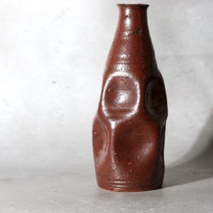 Può includere: Un vaso in ceramica alto e cilindrico con una smaltatura maculata color marrone rossastro scuro. La superficie presenta una serie di profonde rientranze circolari impresse sui lati, creando una forma irregolare e strutturata.