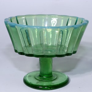 May include: A green glass pedestal bowl with a fluted design. The bowl has a scalloped rim and a round base. The glass has an iridescent quality, with hints of blue and green. The bowl is likely vintage or antique.