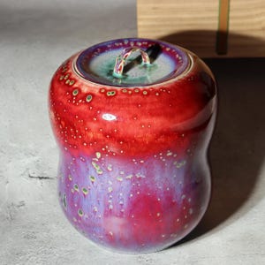 以下が含まれることがあります： 赤から紫への鮮やかなグラデーションが特徴の、蓋付きのセラミックジャー。ジャーは丸みを帯びた形で、蓋と小さなハンドルが付いています。表面には白と緑の点が散りばめられており、デザインにテクスチャを追加しています。