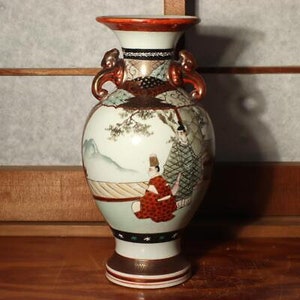 以下が含まれることがあります： 赤と金の縁取りと取っ手のある白い陶器の花瓶。花瓶には、伝統的な日本の服装をした人々と風景が描かれた絵が描かれている。