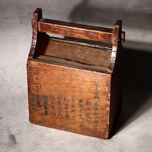 May include: A vintage wooden box with a handle, featuring visible wood grain and dark inscriptions. The box has a rectangular shape with a hinged lid and a sturdy handle for carrying. The box is made of aged wood, showing signs of wear.