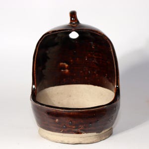 Puede incluir: Un salero de cerámica marrón con forma redondeada y un asa pequeña. El salero tiene una amplia abertura y un interior de color claro. El exterior tiene un acabado brillante con algunas pequeñas imperfecciones.