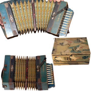 Op de afbeelding: Antieke accordeon met een teal en goud design, met een toetsenbord en balg. De accordeon heeft een leren riem en wordt vergezeld door een bijpassende beschilderde houten kist. De naam van de accordeon is "Garbi".