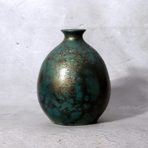 Puede incluir: Un jarrón pequeño y redondeado con cuello estrecho y borde acampanado. La superficie presenta un acabado texturizado y moteado en verde azulado profundo y bronce oscuro, creando un efecto jaspeado con reflejos metálicos en todo el cuerpo.