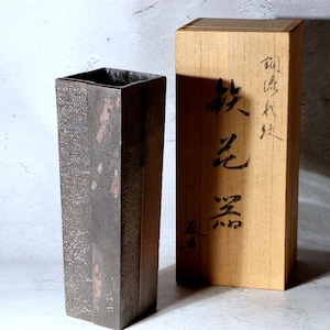 Può includere: Un vaso alto e rettangolare con una superficie strutturata grigio scuro e sottili accenti rosa-marrone. Accanto, una scatola di legno con caratteri giapponesi. Il vaso ha un'apertura quadrata e un design minimalista.