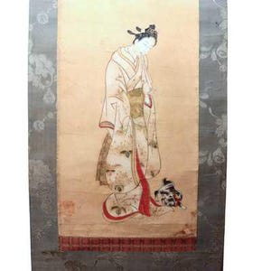 Op de afbeelding: Een verticale hangende rolschildering op een lichtbeige achtergrond met een figuur in een kimono met patronen en rode accenten. Het figuur heeft donker haar dat omhoog is gestyled en wordt vergezeld door een klein donker dier bij de zoom.
