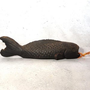 May include: A dark brown, fish-shaped doorstop. The doorstop is made of a solid material, with detailed scales and fins. A small orange rope is attached to the fish's mouth. The doorstop is designed to hold a door open.