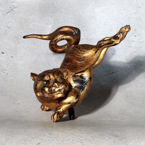 Puede incluir: Una figura de tigre de color dorado en una pose dinámica y saltarina. La escultura presenta un acabado desgastado con acentos negros visibles, rasgos faciales detallados y una cola enroscada. El tigre está capturado en pleno movimiento.