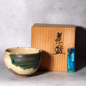May include: A ceramic tea bowl with a cream-coloured crackle glaze and a dark green drip pattern, positioned next to a wooden storage box with black Japanese calligraphy and a blue plastic lighter for scale.