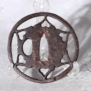 Op de afbeelding: Een cirkelvormige, antieke ijzeren tsuba, een Japanse zwaardbeschermer, met een ingewikkeld opengewerkt ontwerp. De beschermer heeft een centrale opening en decoratieve uitsparingen, met een verweerde, roestbruine patina. De tsuba wordt op een transparante standaard gepresenteerd.