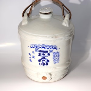 Puede incluir: Una jarra de sake de cerámica blanca con un asa tejida y un pequeño tapón de madera. La jarra presenta caracteres japoneses azules y patrones decorativos. El diseño incluye una boquilla y una tapa con un diseño floral.