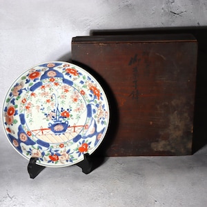 May include: A decorative ceramic plate with a floral design in shades of blue, orange, and white, displayed on a black stand. The plate features a central floral arrangement and a blue border. A wooden box is in the background.
