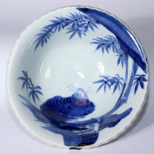 Puede incluir: Un cuenco de porcelana azul y blanco con un diseño de tigre y bambú. El cuenco tiene un borde festoneado y está decorado con un tigre tumbado en una rama de bambú. El tigre está pintado en azul y blanco, y el bambú está pintado en azul.