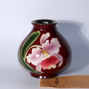 以下が含まれることがあります： 銀色の縁と底が付いた、マルーン色のセラミック花瓶。花瓶には、ピンクと赤の花びらと緑の葉を持つ、描かれた蘭が描かれています。花瓶は丸みを帯びた形状で、滑らかで光沢のある仕上がりです。