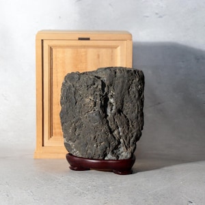 Peut inclure: Une pierre gris foncé à la texture rugueuse et à la surface irrégulière et escarpée est posée verticalement sur un support de présentation en bois brun rougeâtre poli. Derrière la pierre se trouve une boîte en bois rectangulaire claire.