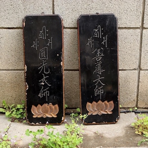 May include: Two rectangular, black wooden plaques with gold-colored trim. Each plaque features carved Asian characters and a lotus flower design. The plaques are weathered and show signs of age, adding to their vintage appeal.