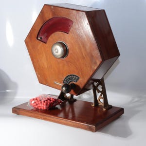 May include: A hexagonal wooden lottery drum mounted on a rectangular base with a metal bell, a red plastic viewing window, and a black handle. A small plastic bag containing numerous small red spherical tokens sits on the base next to the drum.