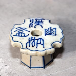 May include: A white ceramic object with blue Chinese characters. The object has a circular hole in the center and a scalloped edge. The base is a white octagon with blue vertical lines. The object is on a gray surface.
