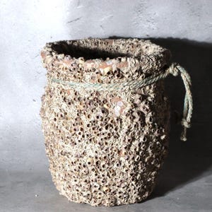 May include: A cylindrical terracotta vessel with a heavily textured exterior covered in numerous small, calcified barnacle-like formations. A weathered, light green rope is tied around the upper rim, featuring a knotted end hanging off the side.