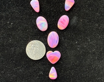 Puede incluir: Una colección de piedras preciosas de ópalo pulidas en varias formas, incluyendo ovaladas, en forma de lágrima y de corazón. Los ópalos muestran una mezcla de tonos rosas, morados y anaranjados. Se incluye una moneda para referencia de tamaño.