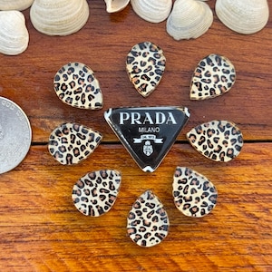 Puede incluir: Una colección de adornos con forma de lágrima con estampado de leopardo y un emblema del logotipo de Prada Milano. Los adornos están dispuestos alrededor del logotipo de Prada sobre una superficie de madera, con conchas marinas esparcidas.