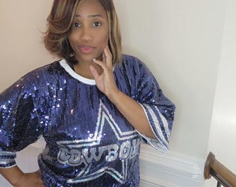 May include: A navy blue sequined short-sleeve shirt featuring a large silver star graphic on the front with the word COWBOYS printed inside the star. The shirt has a white crew neckline and white stripes on the sleeves, creating a sporty aesthetic.