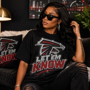 May include: A black short-sleeved t-shirt featuring a rhinestone-embellished graphic of a falcon logo above the text LET EM KNOW in red and silver crystals. The design is repeated on a matching black square throw pillow positioned to the left.