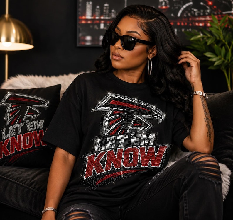 May include: A black short-sleeved t-shirt featuring a rhinestone-embellished graphic of a falcon logo above the text LET EM KNOW in red and silver crystals. The design is repeated on a matching black square throw pillow positioned to the left.