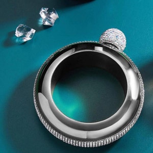 May include: A silver bracelet with a round, clear, rhinestone-covered top. The bracelet is decorated with a row of small rhinestones around the edge.