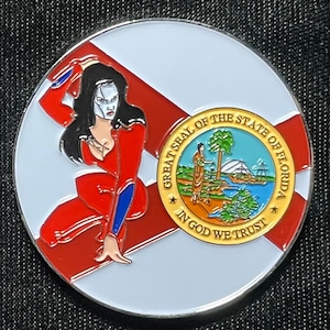 May include: A round, white, metal pin with a red and white Florida state flag design. The flag features a woman in a red and blue suit, and the state seal with the words "Great Seal of the State of Florida" and "In God We Trust".