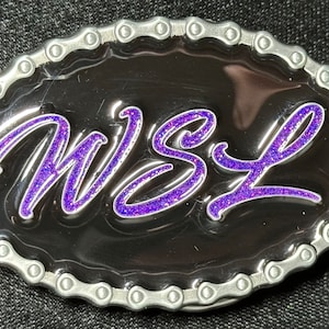 May include: A silver belt buckle with a black background and a purple glitter design that says "WSL".