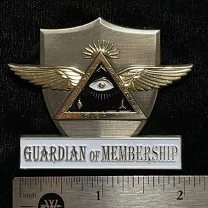 May include: A metal emblem with a shield shape, featuring a triangle with an eye, wings, and the text "GUARDIAN OF MEMBERSHIP" on a white bar. The emblem is approximately 5 cm wide.