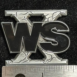 May include: A metal emblem featuring the letters "WS" in black, set against a textured, silver background. The emblem is approximately 5 cm wide, with a cracked, weathered design. The letters are bold and stand out.
