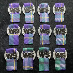 Peut inclure: Douze épingles décoratives avec un motif écossais tissé dans des tons de violet, vert et jaune. Chaque épingle comporte une base circulaire argentée avec les lettres "WS" en noir. Les épingles sont disposées sur une surface noire.