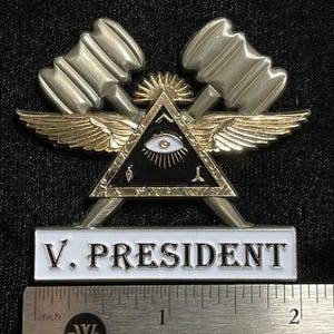 May include: A silver and gold metal pin featuring a triangle with an eye, wings, and two hammers. The pin has the text "V. PRESIDENT" on a white background. The pin is approximately 2 inches long.