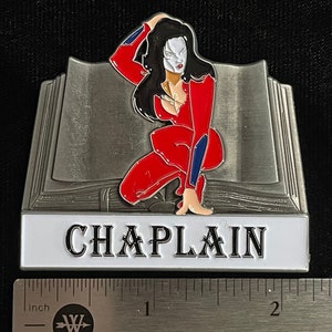 May include: A silver metal pin featuring a woman in a red jumpsuit with a white mask, kneeling on an open book. The book is silver with a white background and the word "CHAPLAIN" in black lettering.