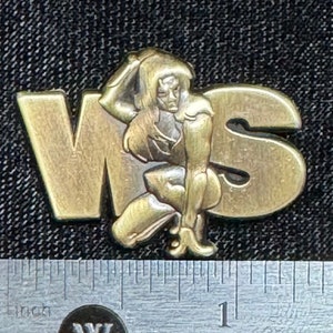 May include: A gold-coloured metal pin with the letters "WS" in raised lettering. A stylised female figure is positioned between the letters.