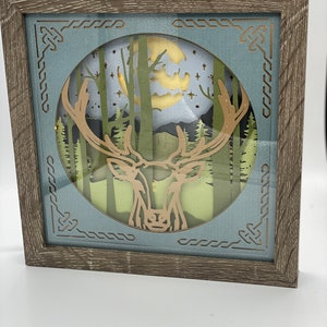 May include: A framed 3D paper art piece depicting a deer with antlers standing in a forest with a full moon in the background. The frame is made of wood with a light brown finish.