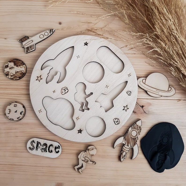May include: A wooden space-themed puzzle with a circular base and various cut-out shapes, including a rocket, astronaut, planet, and stars. The puzzle pieces are made of wood and have a natural finish. The word "SPACE" is written on a separate wooden piece.
