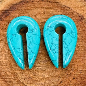 Puede incluir: Un par de expansores de oreja de color turquesa en forma de lágrima con un agujero central. Los expansores tienen un patrón de mármol.