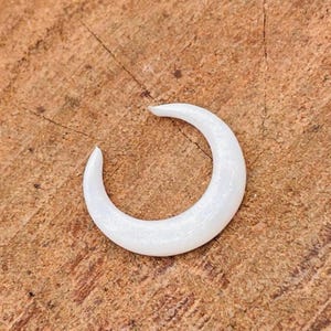 Puede incluir: Un pendiente blanco en forma de media luna. El pendiente tiene un diseño liso y curvado y un acabado brillante. El pendiente está sobre una superficie de madera.