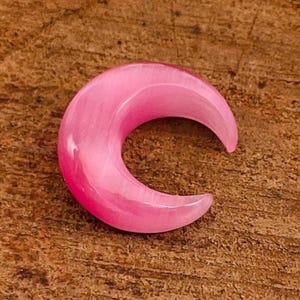May include: A pink, crescent-shaped ear gauge plug. The plug is made of a translucent material, with a smooth, polished surface. The color is a vibrant, light pink. The background is a textured, brown surface.