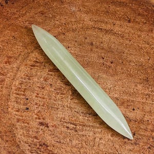 Puede incluir: Una talla de jade verde claro en forma de hoja larga y delgada. La hoja es lisa y pulida, con una punta ligeramente redondeada.