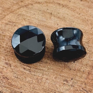 May include: Two black ear gauges, one with a faceted design and the other with a smooth, rounded surface.