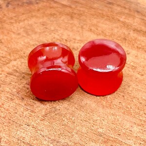 May include: Two red acrylic ear gauges, one is a flared style and the other is a simple cylinder.