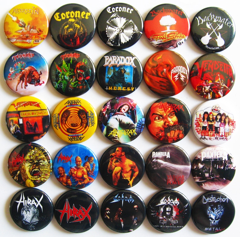 May include: A collection of 20 round metal pinback buttons featuring various heavy metal band logos and album art. The buttons are black, yellow, red, blue, and white with images of skulls, band names, and album titles. The bands featured include Slayer, Anthrax, Coroner, Paradox, and Pantera.