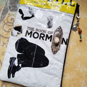 Puede incluir: Una bolsa blanca con cremallera y diseño acolchado. La bolsa presenta una imagen en blanco y negro de una persona arrodillada con un micrófono y el texto "PLAYBILL EUGENE O'NEILL THEATRE THE BOOK OF MORMON".