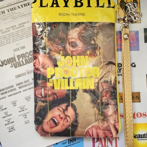 May include: A rectangular zippered pouch featuring a Playbill cover design for the production John Proctor Is The Villain at the Booth Theatre. The pouch has a yellow top border, a photographic print of laughing faces, and a gold wrist strap attached.