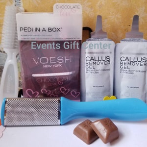 May include: A pink and purple box with the text "PEDI IN A BOX&reg;" and "Events Gift Center" on it. The box contains a foot scrub, callus remover gel, and chocolate. The box is on a white surface.